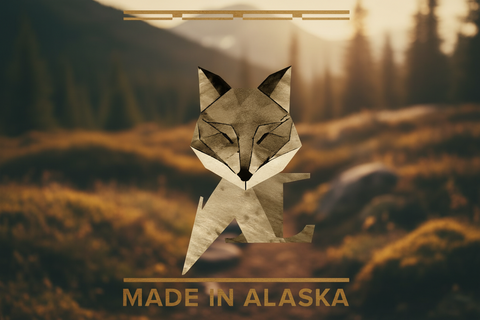 A stylized low-poly fox graphic centered over a blurred mountain landscape with trees and warm golden light. The text 'MADE IN ALASKA' appears at the bottom in a gold-toned font, with matching horizontal lines near the top.