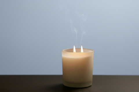 A lit white candle in a frosted glass holder, pale blue and brown background. Gentle smoke rises from the candle.