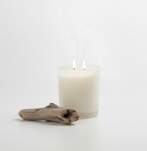 A lit white candle in a frosted glass jar with two wicks, placed on a white surface beside a small piece of driftwood against a clean white background.