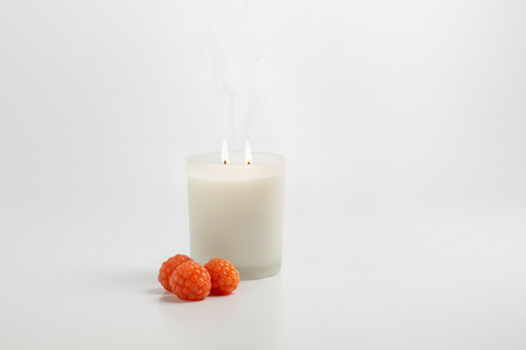 A white wax candle in a frosted clear glass jar with two lit wicks and visible rising smoke, styled with three orange salmonberries on a clean white surface against a white background.