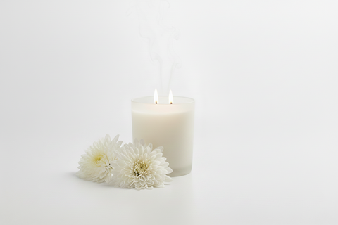 candle with two wicks burning in a clean white studio setting.   two white mum flowers sit to the left side of the candle.