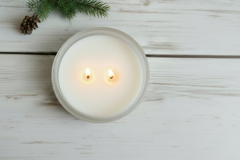 A lit white candle in a round clear glass jar sits on a white wooden surface, with pine branches and a small pinecone placed in the upper left corner.