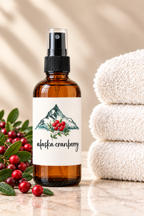 An amber glass spray bottle labeled 'alaska cranberry' with a black spray pump, set on a marble surface with red cranberries and green leaves on the left and a stack of white towels on the right.