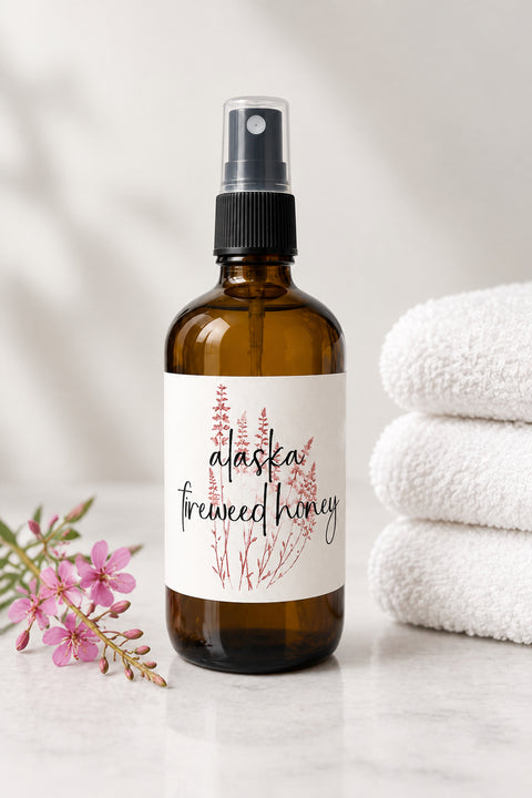 An amber glass spray bottle labeled 'alaska treeweed honey' with a black spray top, styled on a light marble surface with pink wildflowers on the left and a stack of white towels on the right.