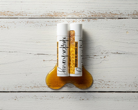 Two lip balm tubes labeled alaska honey balm and a design of honeycomb with a hexagon pattern, placed on top of a pool of golden honey on a white rustic wooden background.