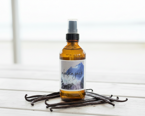 Alaska Jack linen spray and room mist in amber glass bottle with black pump top, styled on a white wood surface surrounded by vanilla bean pods with soft coastal light in the background, Grey Fox Candles