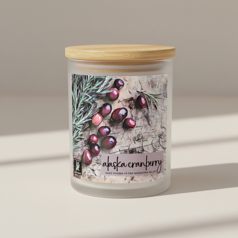Alaska cranberry candle label artwork showing glossy dark red cranberries scattered on a textured stone surface with green pine sprigs. Text reads 'alaska cranberry' and 'HAND POURED IN THE MATANUSKA VALLEY, grey fox candles.