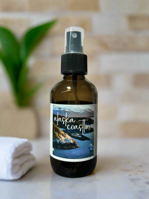Brown spray bottle with 'Alaska Coastline' label on a marble surface with a plant and towel in the background.