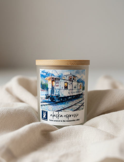 Alaska Espresso coconut wax candle in white frosted vessel with bamboo lid, styled on a warm wooden dining table next to a steaming coffee cup and eucalyptus sprig, Grey Fox Candles