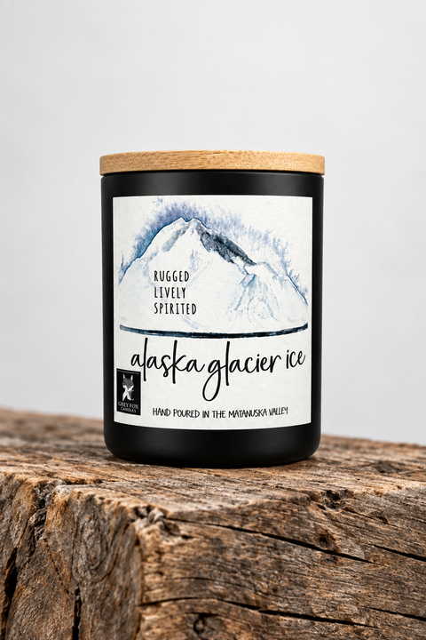 A matte black glass candle jar with a bamboo lid sits on a rustic wood beam against a plain light gray background. The front label reads 'alaska glacier ice' with a blue-and-white mountain illustration and smaller text 'Rugged, Lively, Spirited.