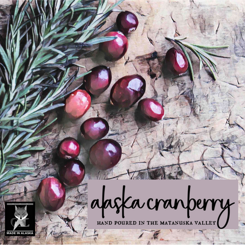 Alaska cranberry product label artwork showing glossy dark red cranberries scattered on a textured stone surface with green pine sprigs. Text reads 'alaska cranberry' and 'HAND POURED IN THE MATANUSKA VALLEY, grey fox candles.