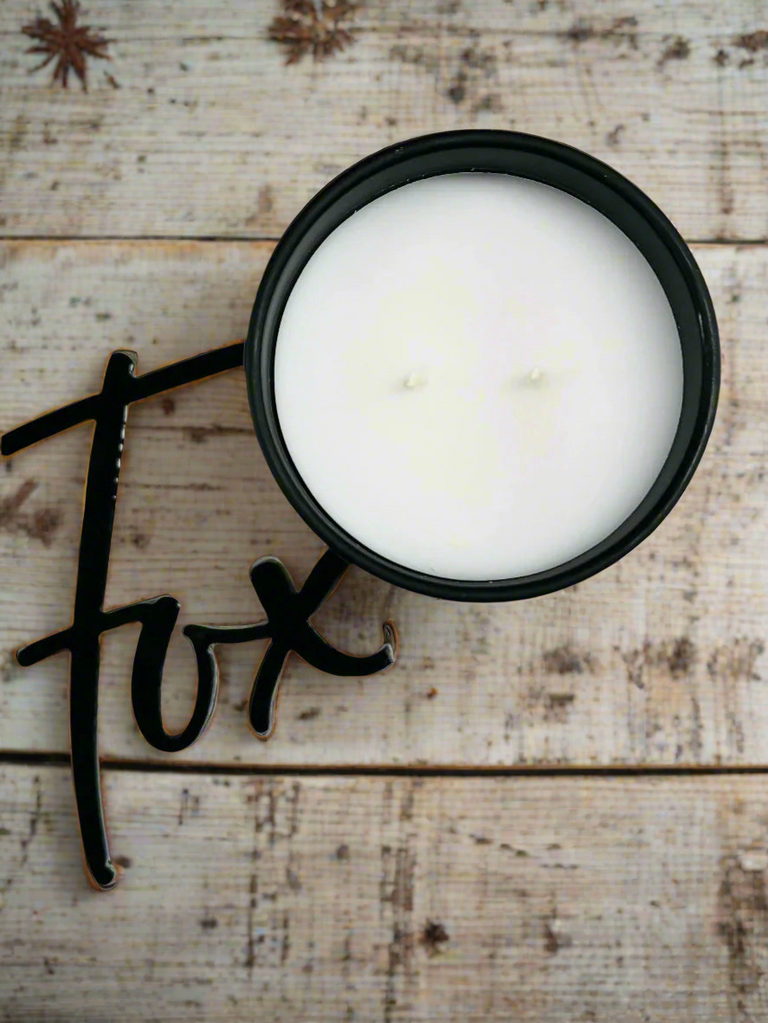 White candle in a black container on a wooden surface with the word 'fox' written in black.