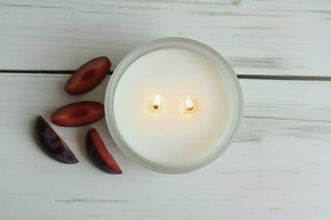 A lit white candle in a clear glass jar sits on a white wooden surface next to sliced dark purple plums.