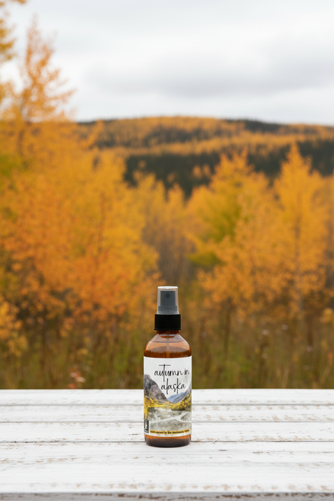 An amber glass spray bottle with a black pump dispenser and a white label featuring a landscape illustration of autumn in Alaska