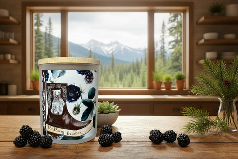 Candle with a blackberry bourbon label on a wooden table with blackberries, plants, and a mountain view.