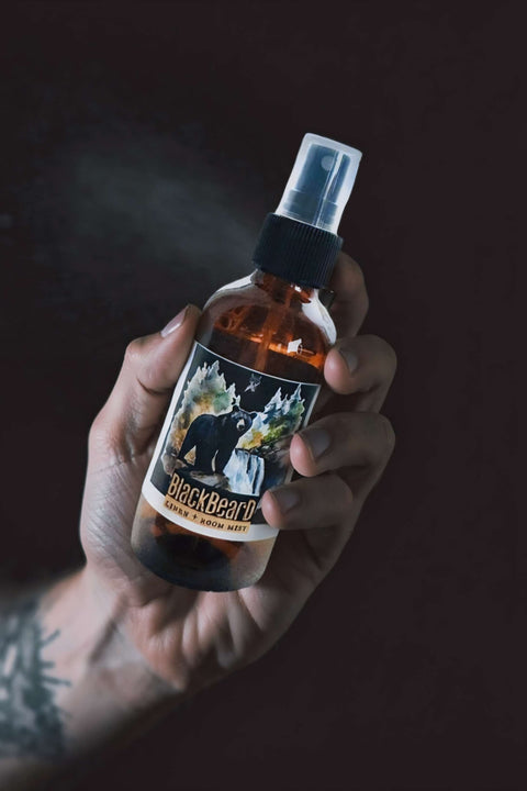 A hand holding an amber glass spray bottle with a black spray top and a label reading BlackBear. The bottle is shown against a very dark, near-black background with a faint mist or smoke effect around it.