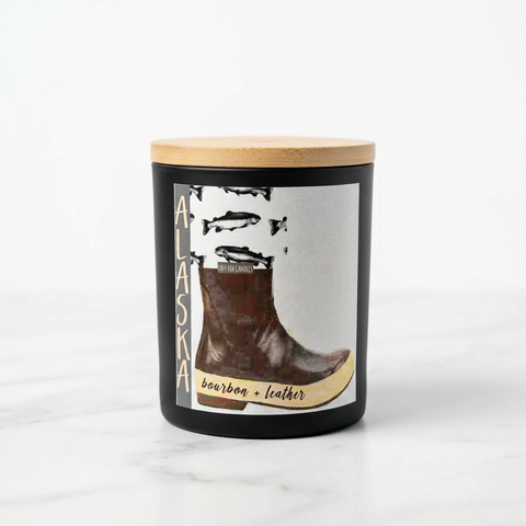 A black jar candle with a light wood lid, featuring a graphic label of a brown leather boot and the text 'ALASKA bourbon + leather'.