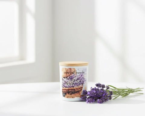 A white glass jar candle with a light wood lid sits on a white surface next to a small bouquet of purple lavender flowers. The candle label features a photo of lavender in a basket with the text 'made in alaska'