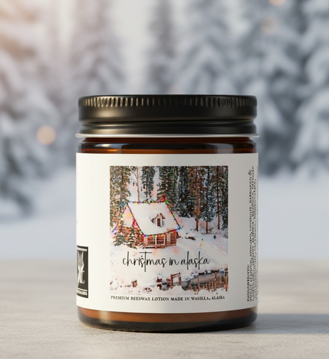 A single amber glass jar candle with a black metal lid and a white label reading 'christmas in alaska,' photographed on a light surface with a blurred snowy outdoor background.