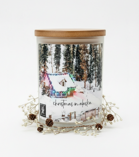 A glass jar candle with a light wood lid and a winter forest label reading 'christmas in alaska,' styled with white berries and pinecones on a white background.
