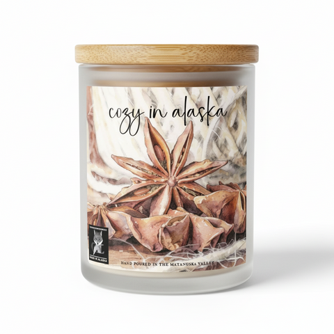 a candle with the words 'cozy in alaska' over a watercolor-style illustration of star anise and dried botanical elements. A small 'Made in Alaska' logo appears in the lower left, with additional text reading 'Hand poured in the Matanuska Valley' at the bottom.