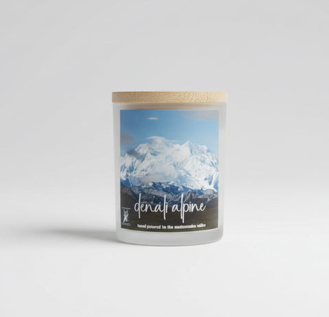Candle with mountain view watercolor label on a white surface with a  Denali mountain in the background with the text 'Denali alpine' and hand poured in the matanuska valley