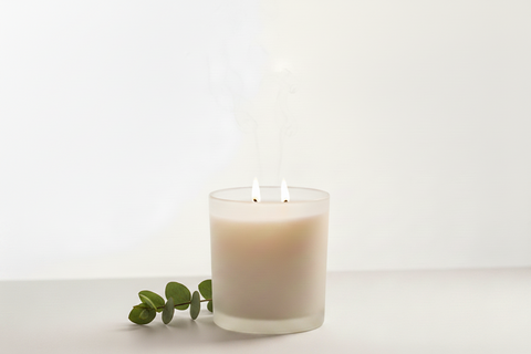 A frosted glass candle holder with two lit wicks and wisps of smoke, set against a clean white background.