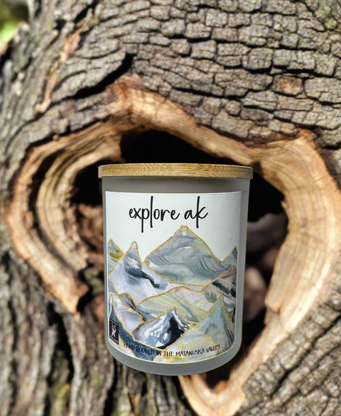 Candle with 'explore ak' text and mountain design in a natural setting