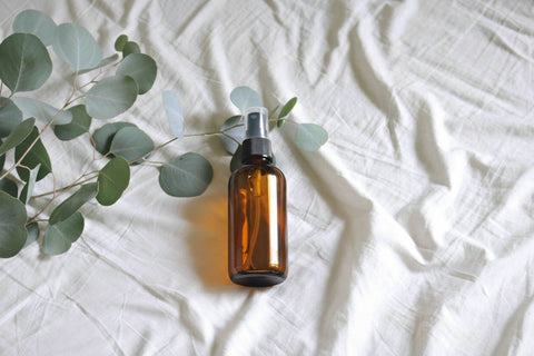 bottle of eucalyptus white tea linen spray on white bedding, fresh eucalyptus plant sprig lying next to it on the bed