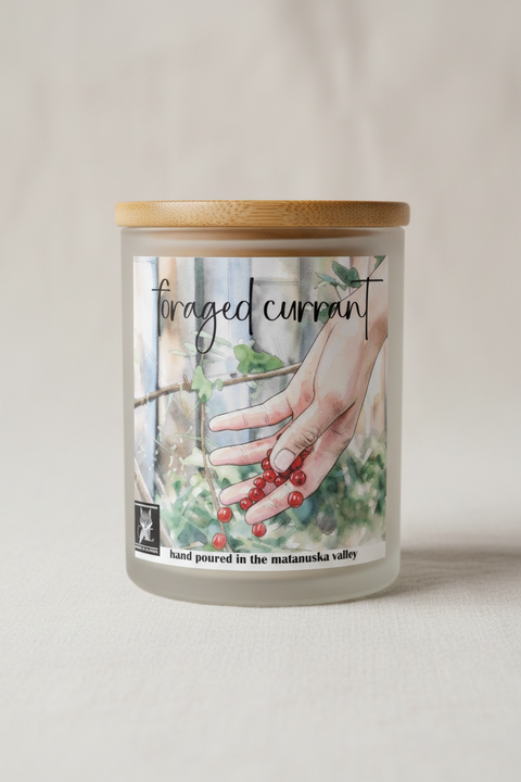 A frosted glass candle jar with a light wood lid, featuring a label that reads 'foraged currant' and shows a hand holding red berries. The candle is photographed on a neutral, light beige textured surface with a soft, warm background.