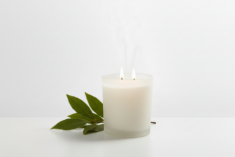 A frosted glass candle holder with two lit wicks and wispy smoke rising, styled with leaves on a clean white surface against a white background.