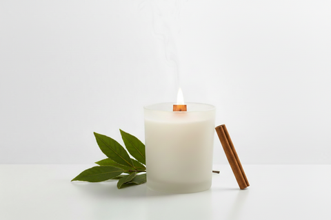 A white scented candle in a frosted glass holder with a wood wick, shown lit with a wisp of smoke rising. Green leaves and a cinnamon stick rest beside it on a clean white surface against a white background.