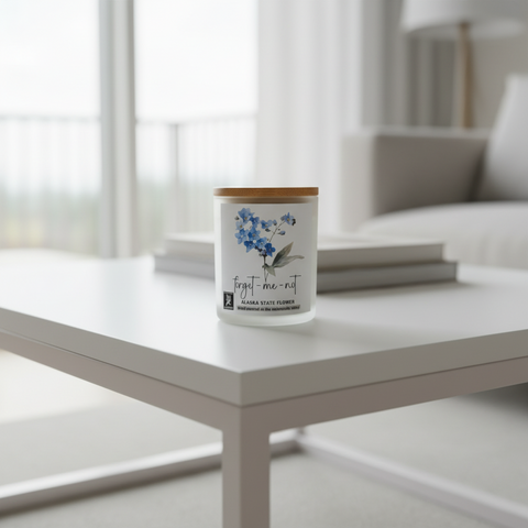 A jar candle labeled 'forget-me-not' with blue flower artwork sits on a white coffee table in a white living room