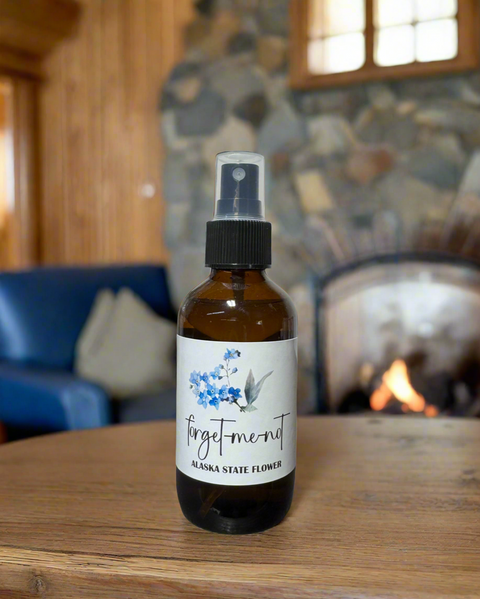 Bottle of 'Forget-me-not' linen and room spray with a label featuring flowers on a wooden surface with a fireplace in the background.