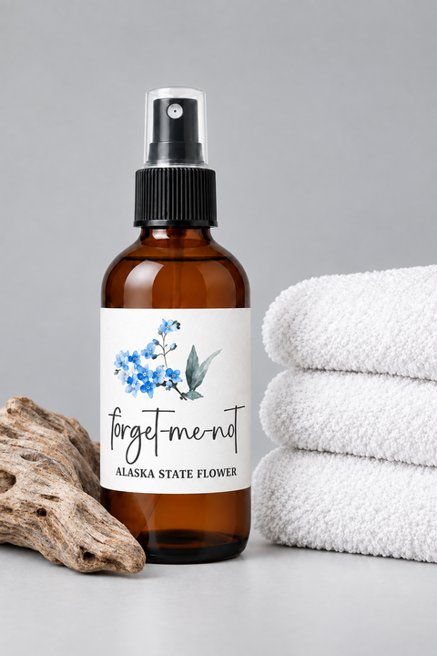 An amber glass spray bottle labeled 'forget-me-not ALASKA STATE FLOWER' with blue forget-me-not flowers printed on the label, styled on a gray surface with white folded towels and a piece of driftwood.