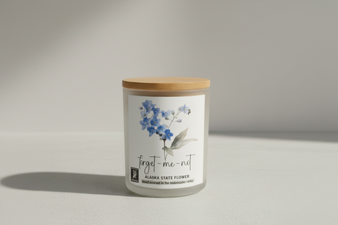 Candle with a floral label on plain surface in a studio setting. text says 'forget me not ' and 'alaska state flower'