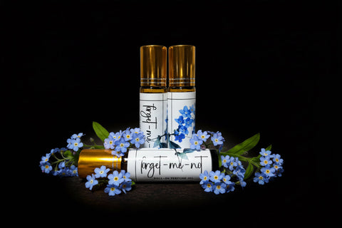 Three small bottles with gold caps and white labels surrounded by forget me not flowers on a black background