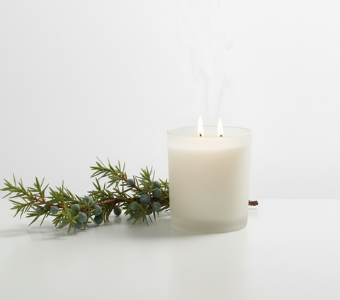 A frosted glass candle with two lit wicks and visible rising smoke, placed on a white surface beside a green sprig with small berries against a clean white background.