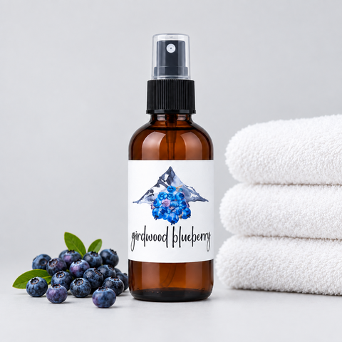 An amber glass spray bottle labeled girdwood blueberry with blueberries and a mountain printed on the label, styled on a gray surface with white folded towels and blueberries scattered.