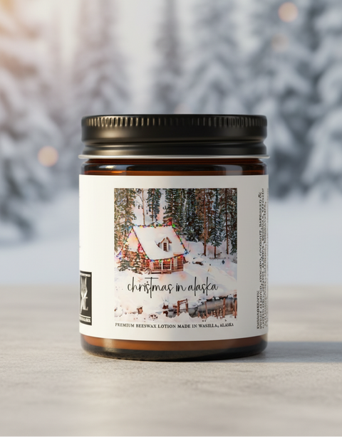 A rectangular product label or packaging panel with a winter cabin scene under colorful string lights, overlaid with the text 'christmas in alaska.' Smaller text at the bottom reads 'PREMIUM BEESWAX LOTION MADE IN WASILLA, ALASKA.