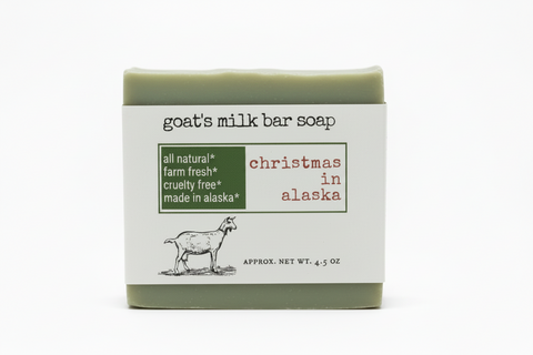 Product label for goat's milk bar soap with the scent 'christmas in alaska'. The label features a green box with white text listing key attributes, red text for the scent name, a black-and-white line drawing of a goat, and the net weight at the bottom.
