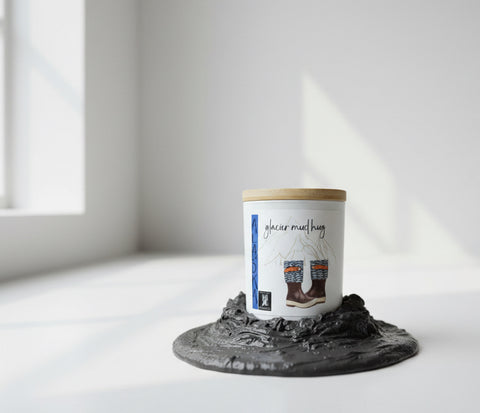 Glacier Mud Hug coconut wax candle in white vessel with bamboo lid, sitting on dark glacier mud in a white studio setting