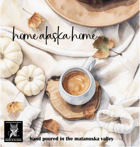 A square watercolor-style product graphic with the words 'home alaska home' across the center and 'hand poured in the matanuska valley' at the bottom. The scene shows a cup of coffee on a wooden coaster, cream knit fabric, small white pumpkins, scattered autumn leaves, and part of a hat, with a small 'Made in Alaska' logo in the lower left.