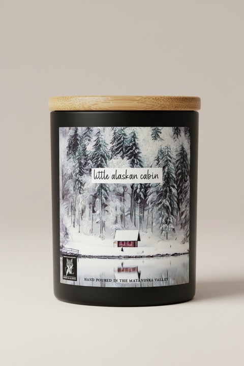 watercolor artwork showing a snowy forest with a little red cabin and a calm lake, overlaid with the text 'little alaskan cabin' and 'HAND POURED IN THE MATANUSKA VALLEY.' A small logo in the lower left reads 'Made in Alaska. by grey fox candles