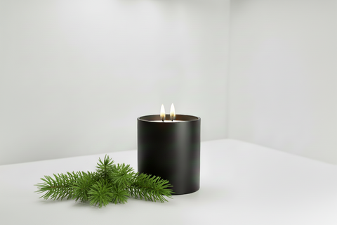 A black cylindrical candle with two lit wicks burning in a studio setting and a pine sprig.