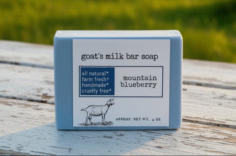 Bar of goat's milk soap labeled 'mountain blueberry' on a wood farmhouse table, with a blurred background of sheep in a field.