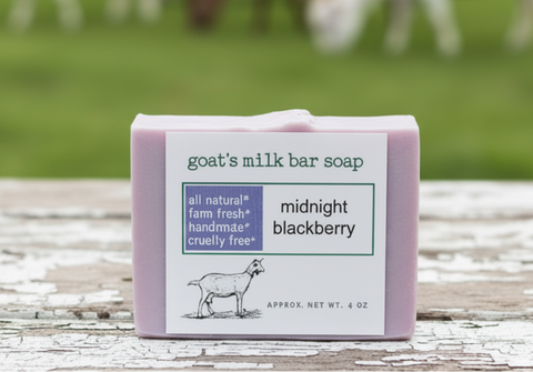 A bar of goat's milk soap labeled 'midnight blackberry' sits on a weathered white wooden table, with a blurred green field and grazing goats in the background.