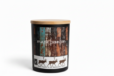 A matte black candle jar labeled 'maple bourbon' sits in a white studio setting. label shows a multi colored plank peeled boards  with 3 caribou along the bottom and text that says ' hand poured in the matanuska valley, made in alaska logo by grey fox candles