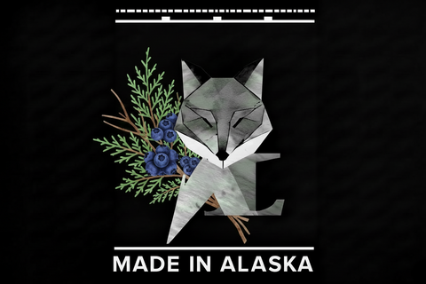 A black graphic design featuring a stylized gray fox head layered with green foliage and blue berries on the left. White text at the bottom reads 'MADE IN ALASKA,' and decorative white lines appear at the top and bottom.