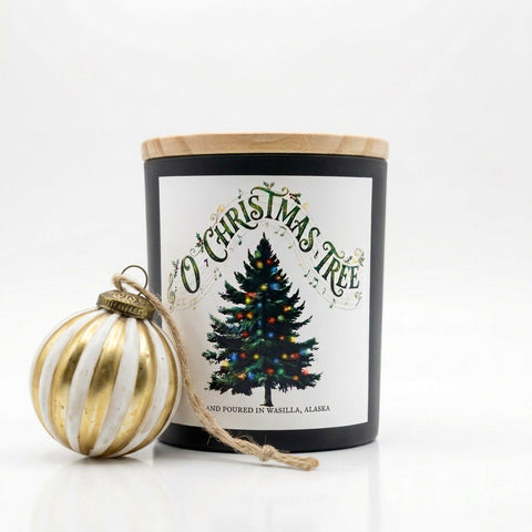 A black glass jar candle with a light wood lid and a white label reading 'O Christmas Tree' with a decorated tree illustration, shown beside a gold-and-white striped ornament on a white background.